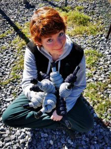 Frau mit kurzem, rötlichem Haar sitzt im Schneidersitz auf Kies und hält mehrere dunkelblaue und silberne Ropeflow-Seile wie einen Blumenstrauß – Symbol für kreative Bewegung und Leichtigkeit.